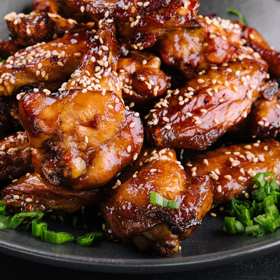 Close-up of glazed chicken wings topped with sesame seeds and chopped green onions on a dark plate.